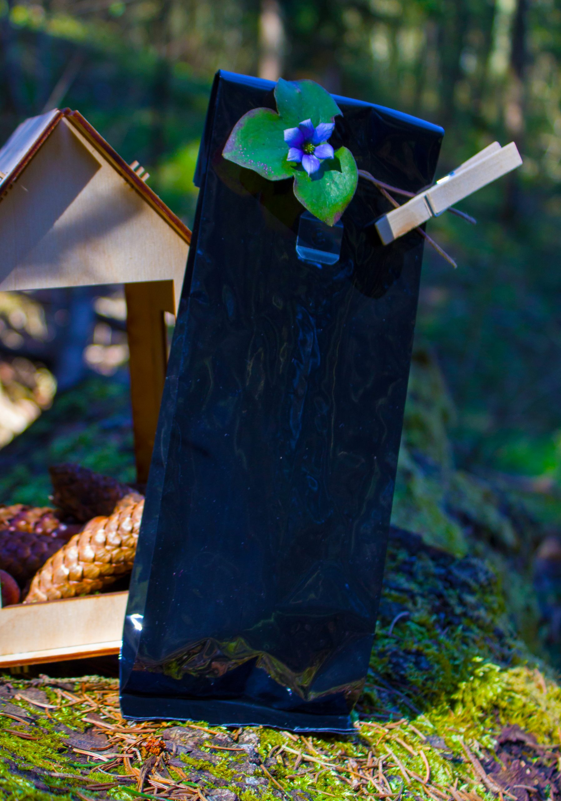 Juodas mobilusis telefonas, pritvirtintas prie medžio kamieno su spaustuku, ant jo uždėta mėlyna gėlė su žaliais lapais, fone matomas medinis paukščių namelis ir spygliuočių kankorėžiai.