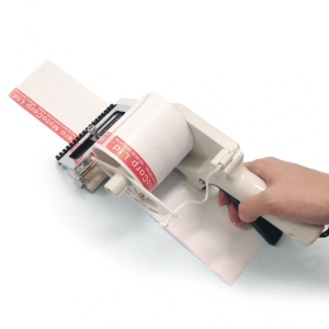 Rankenos laikoma etikečių klijavimo mašinėlė, naudojama lipnioms etiketėms klijuoti ant balto paviršiaus.