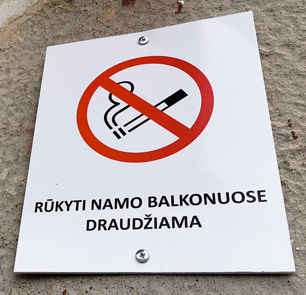Kvadratinėje baltoje lentelėje pavaizduotas rūkymo draudimo simbolis ir po juo užrašas lietuvių kalba 