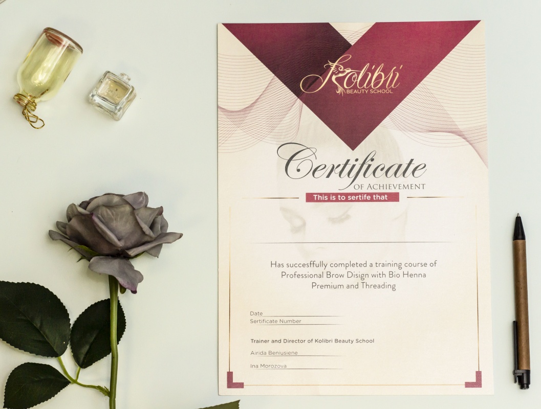 Certifikato dokumentas ant šviesaus paviršiaus su rožinėmis ir bordinėmis detalėmis, šalia džiovintos rožės, rašiklio ir uždaryto stiklinio indelio.