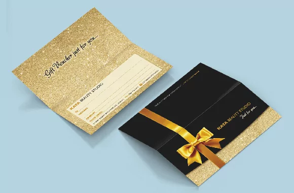 Dvi dovanų čekio kortelės ant melsvos spalvos fono. Viena kortelė yra su blizgančiu auksiniu paviršiumi, o antroji - juoda su auksine kaspinu.